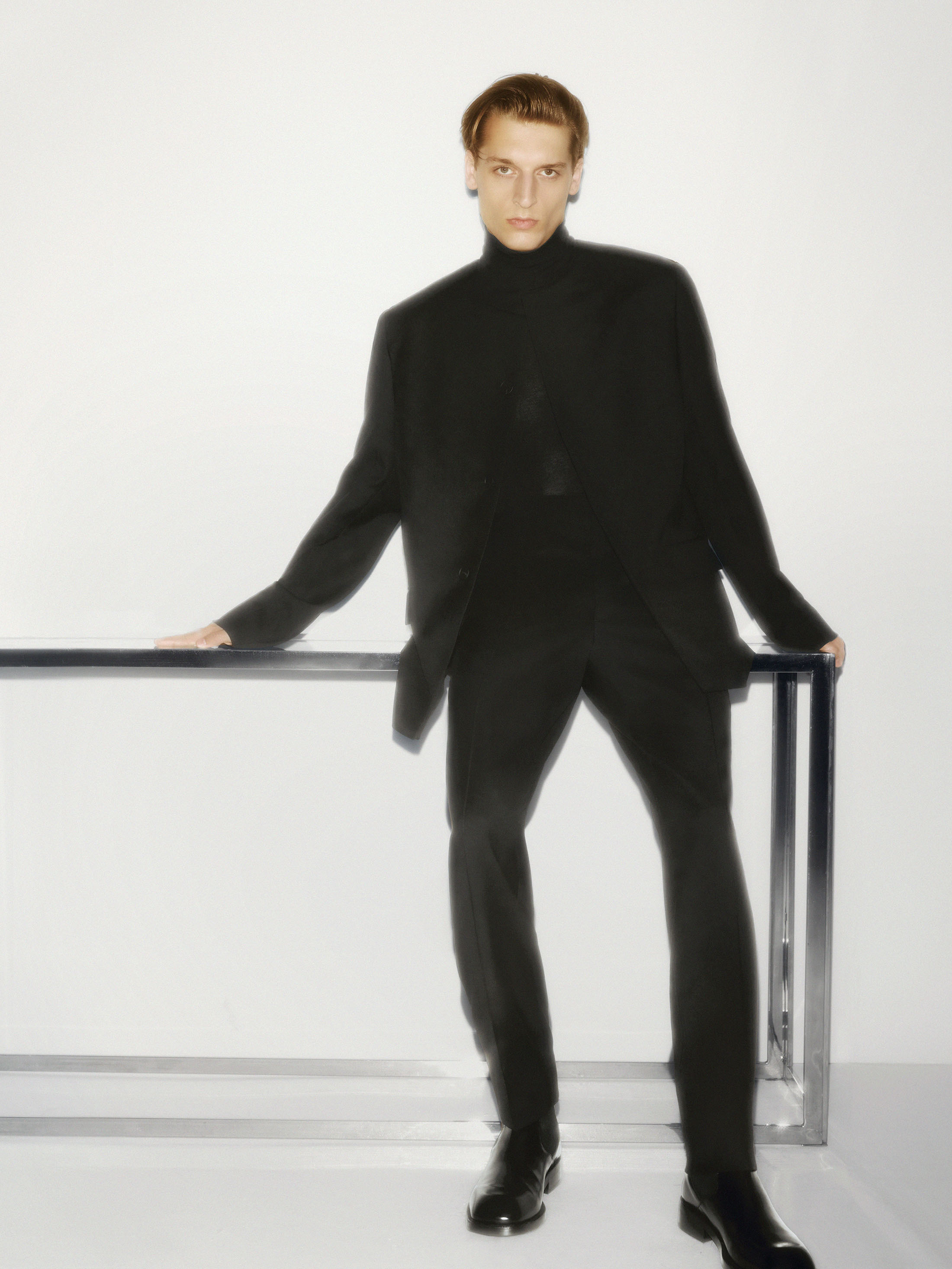 Man wearing a black turtleneck and blazer leaning on a metallic frame against a white studio background.