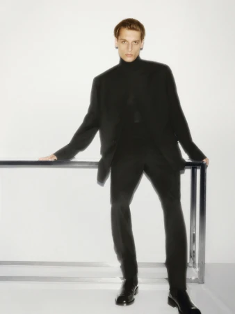 Man wearing a black turtleneck and blazer leaning on a metallic frame against a white studio background.