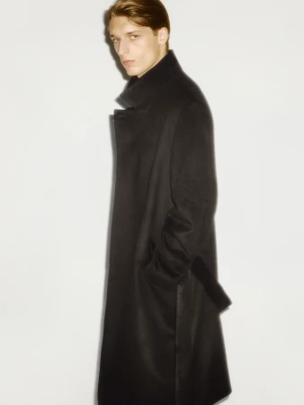 Model wearing a long black coat with a high collar against a plain white studio background.
