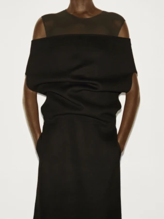 Model wearing a structured black dress against a plain off-white studio background for the COS Fall 2025 campaign.