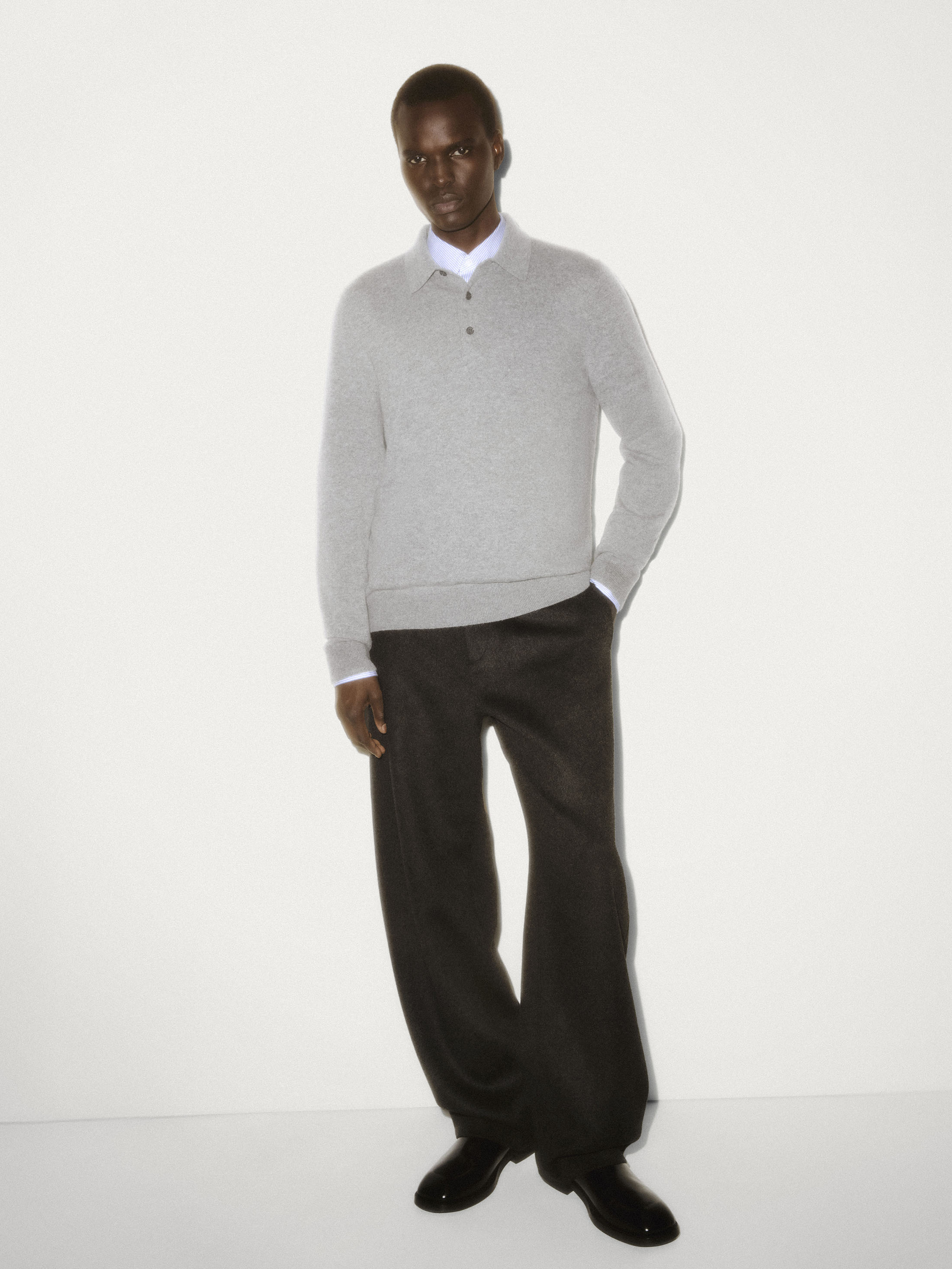 Black man wearing a grey knit polo shirt and dark trousers against a white seamless studio background.
