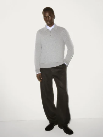 Black man wearing a grey knit polo shirt and dark trousers against a white seamless studio background.