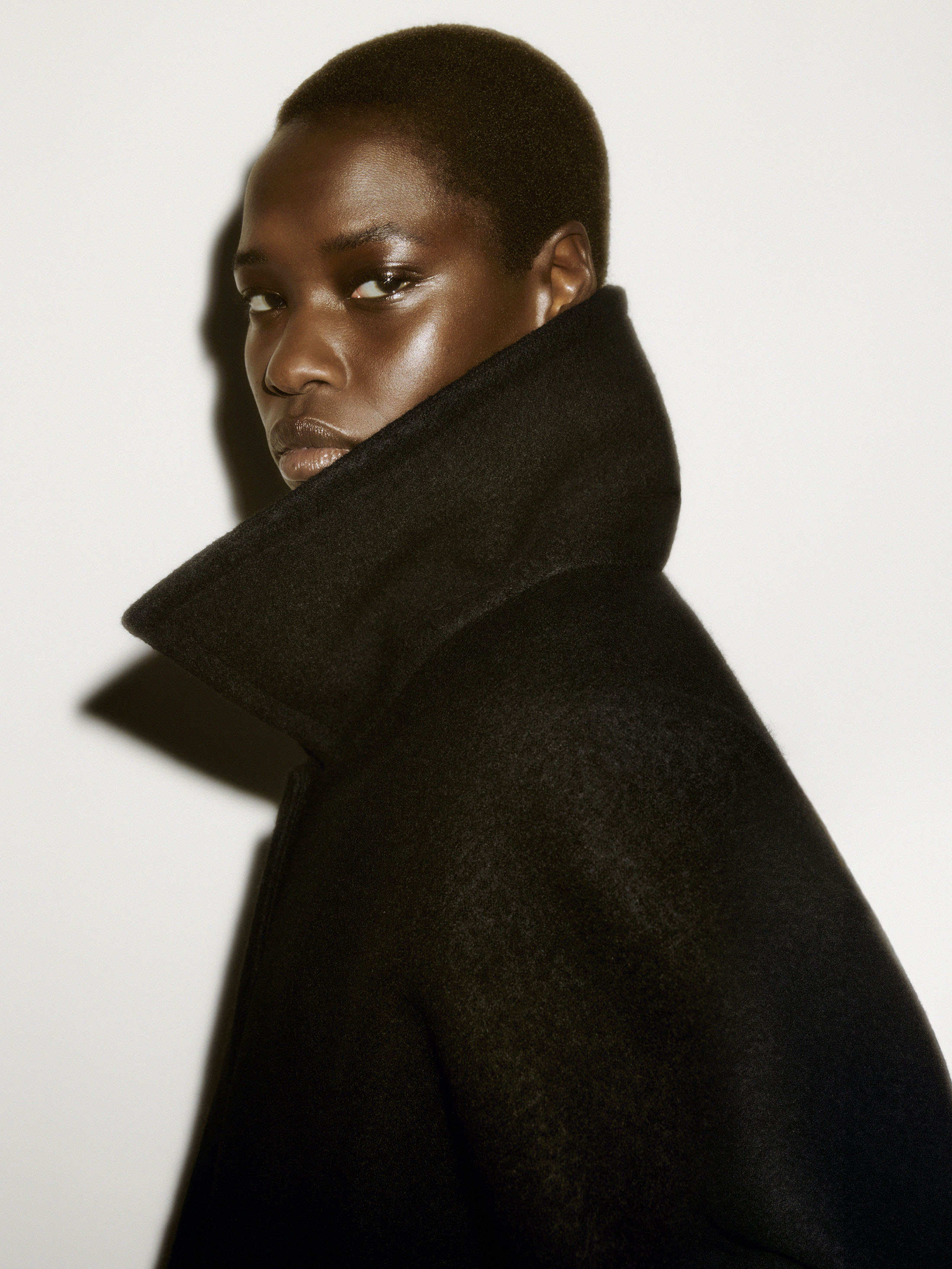 Close-up portrait of a model with short dark hair wearing a high-collared black coat against a plain white studio background.