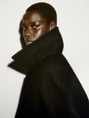 Close-up portrait of a model with short dark hair wearing a high-collared black coat against a plain white studio background.