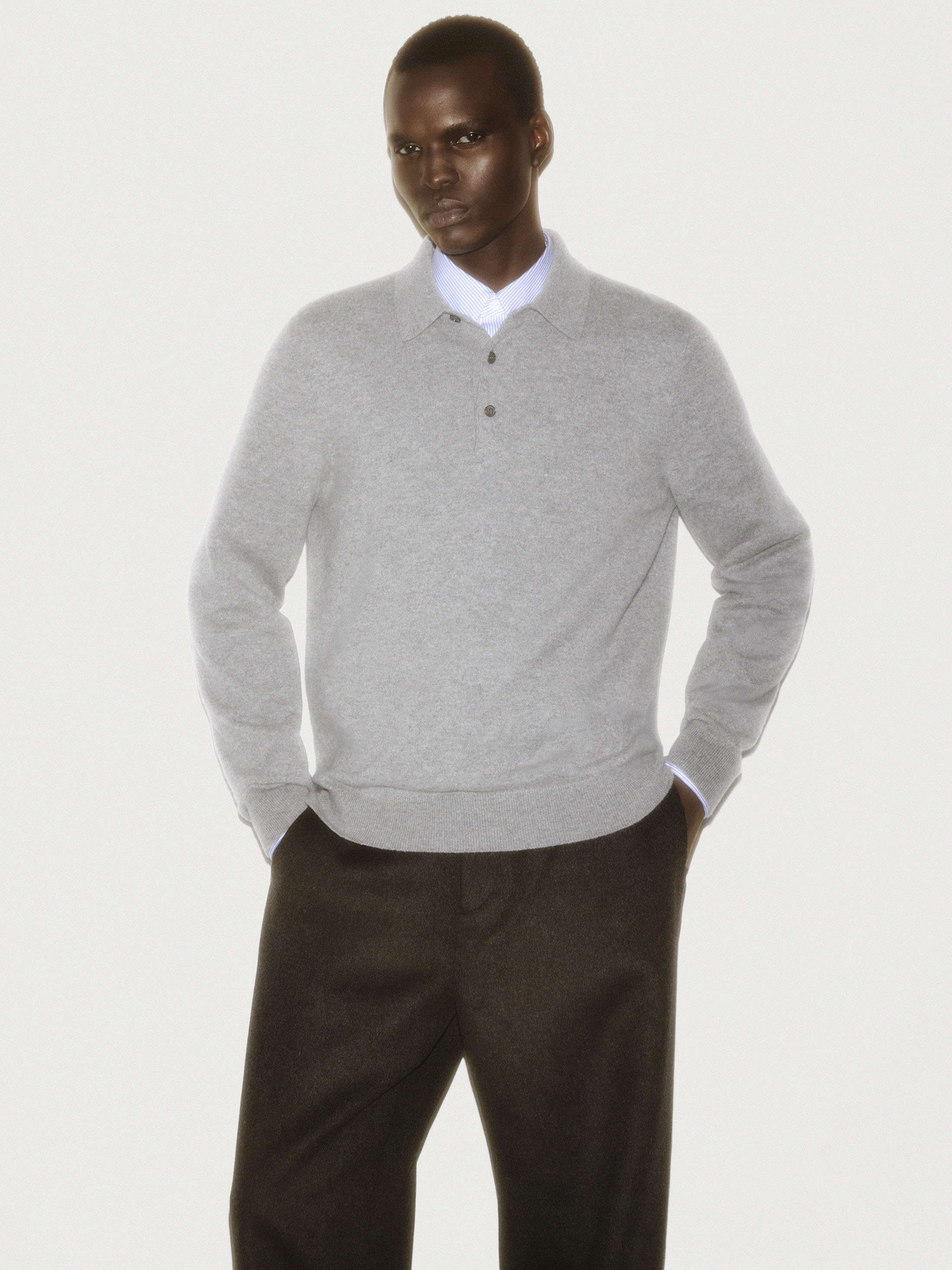 Man wearing a grey polo sweater and dark trousers against a white background for a COS ad campaign