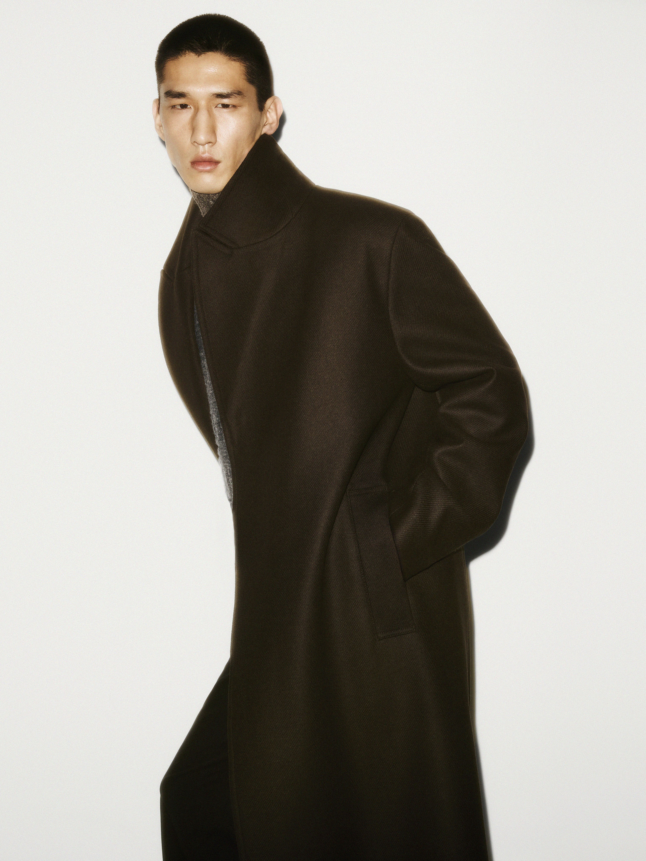 Man wearing a long dark brown structured coat standing against a plain white background for COS