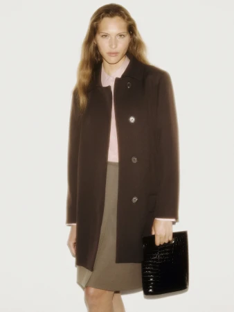 Model in brown coat wearing pink shirt and holding a black handbag against a plain background