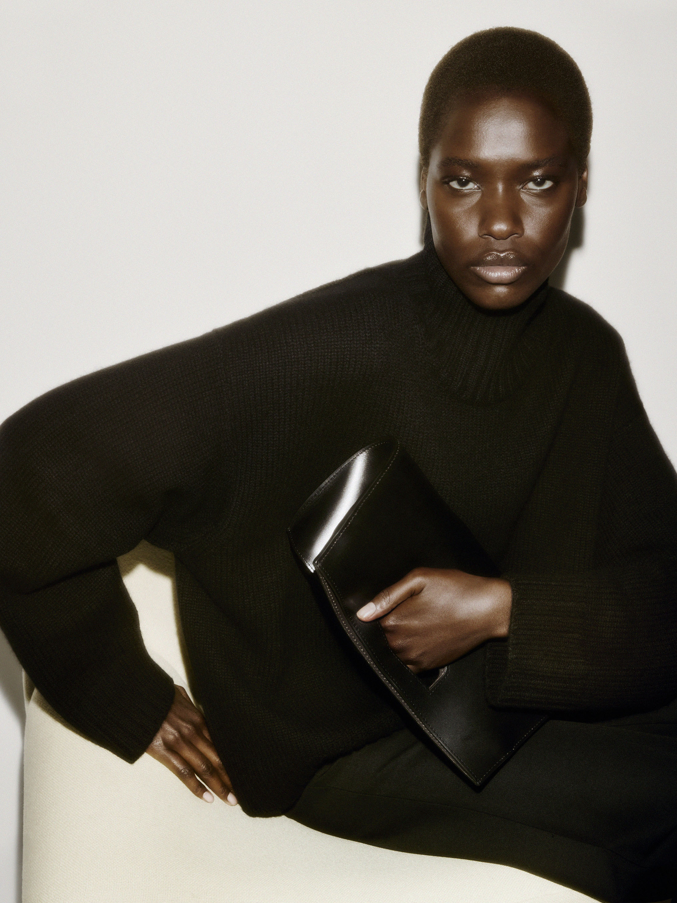 Model wearing a dark knit sweater holding a black leather clutch against a pale backdrop
