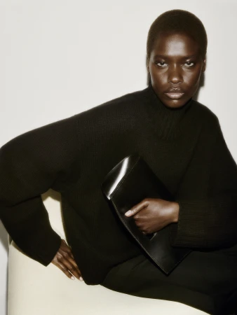 Model wearing a dark knit sweater holding a black leather clutch against a pale backdrop