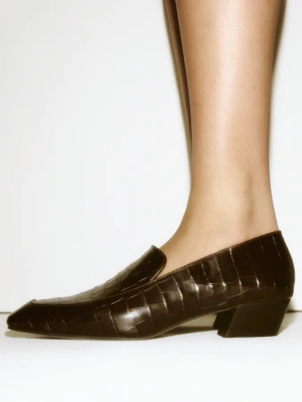 Close-up profile of a person wearing black embossed leather loafers against a plain white studio background for COS Fall 2025 ad campaign.