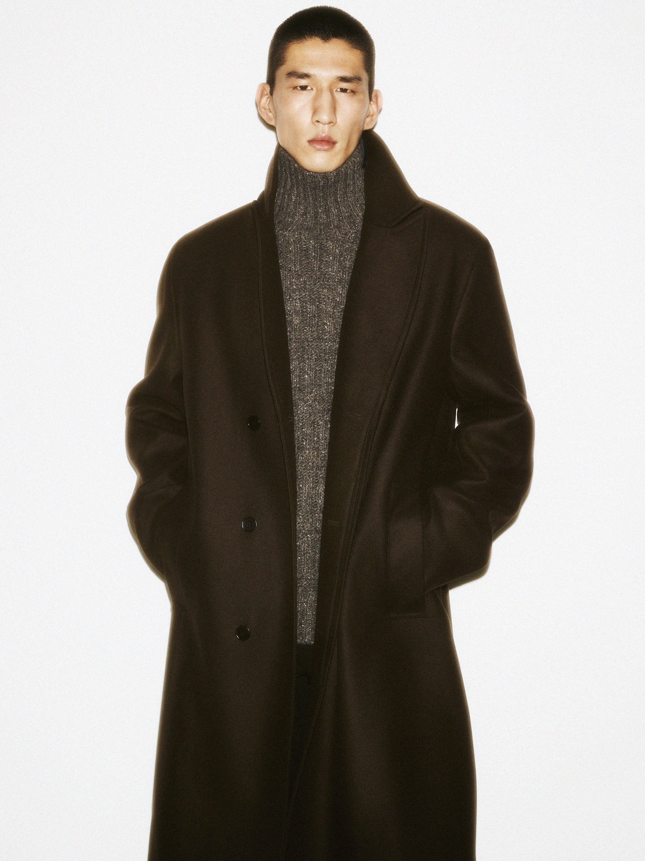 Man in a dark coat and grey turtleneck sweater standing against a clean white background