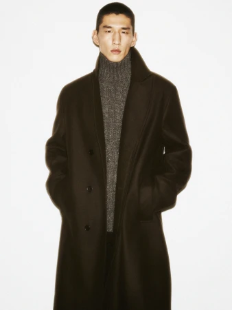 Man in a dark coat and grey turtleneck sweater standing against a clean white background