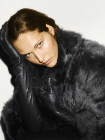 Model in dark faux fur coat and long leather gloves against a plain background