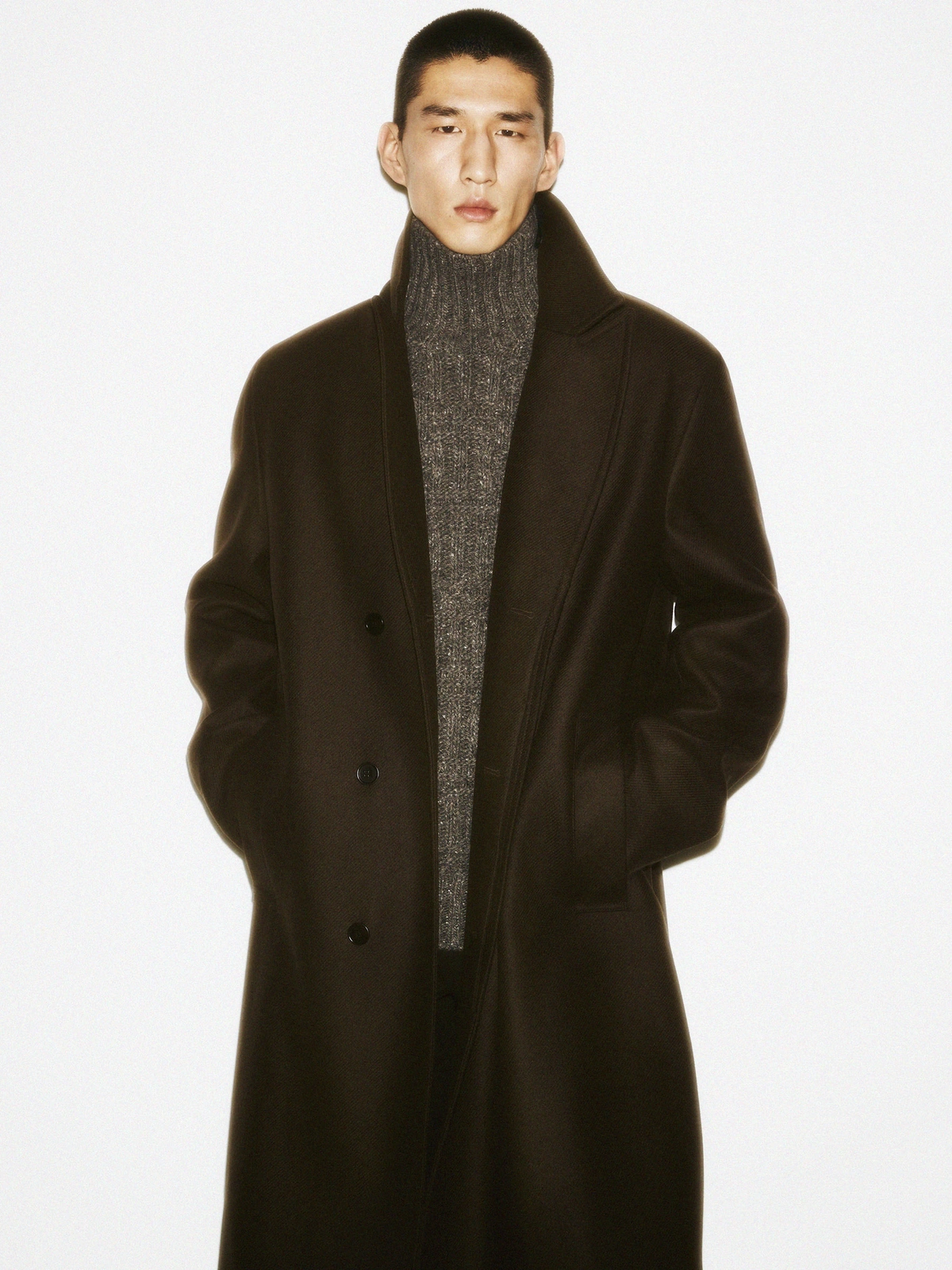 Man in a dark coat and grey turtleneck sweater standing against a clean white background