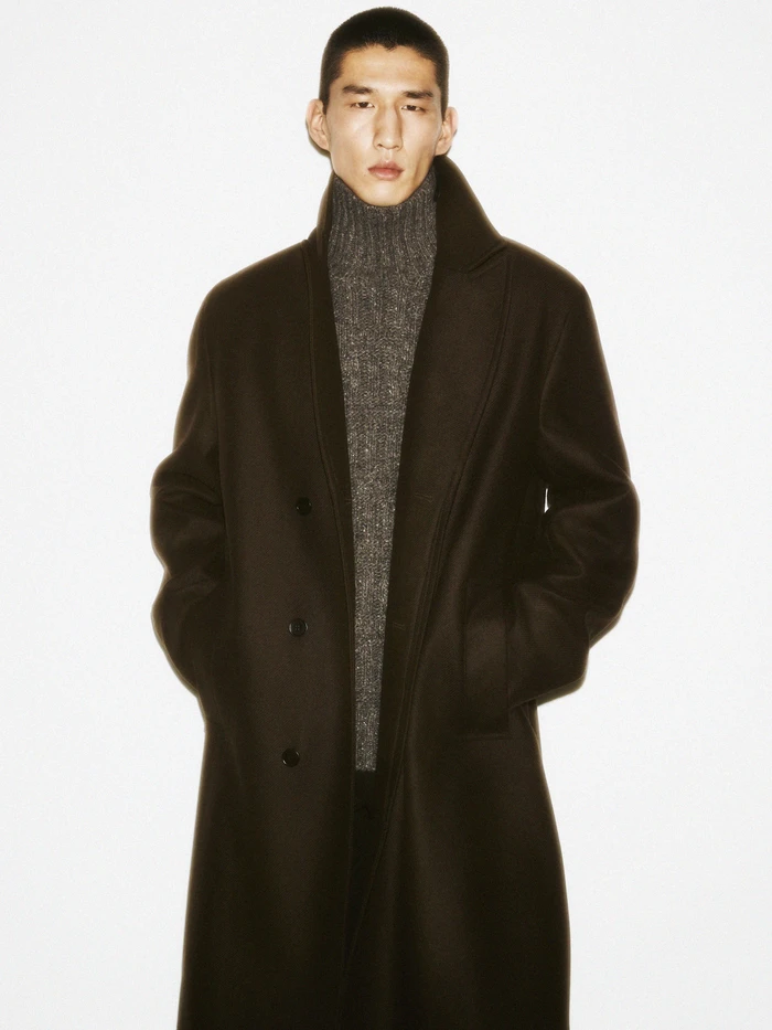 Man in a dark coat and grey turtleneck sweater standing against a clean white background