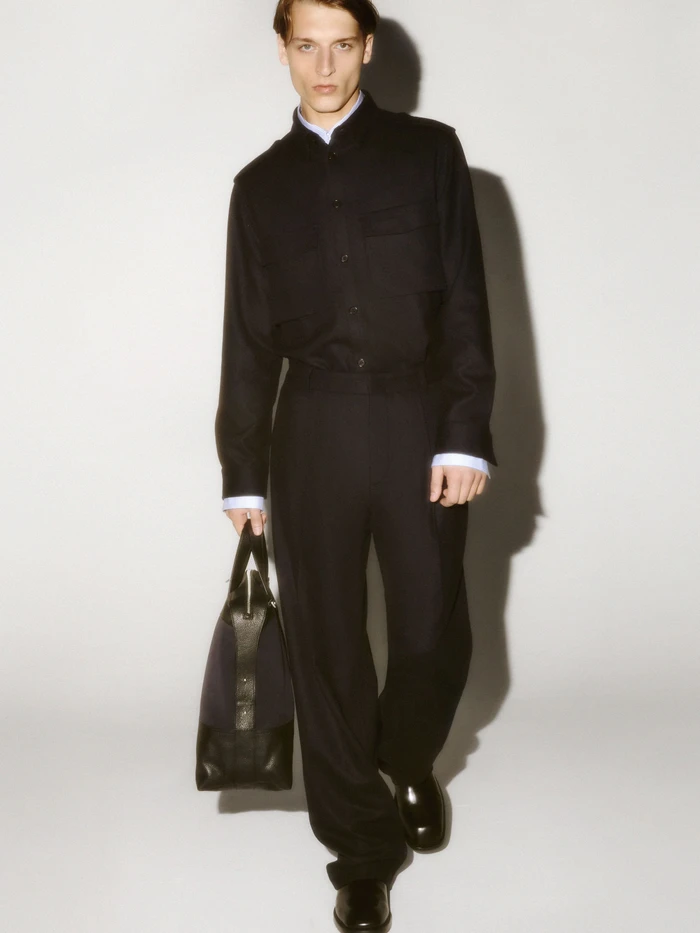 Man wearing a dark button-up shirt and trousers holding a black handbag against a seamless white background