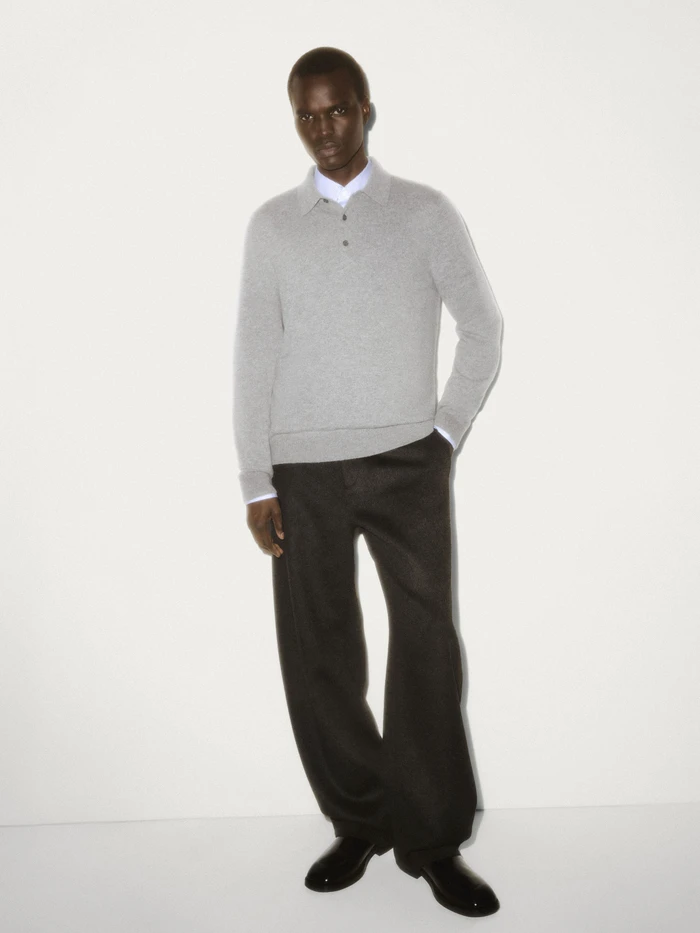 Black man wearing a grey knit polo shirt and dark trousers against a white seamless studio background.