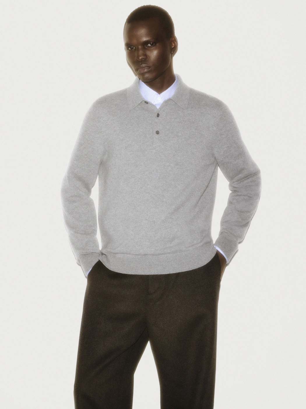 Man wearing a grey polo sweater and dark trousers against a white background for a COS ad campaign