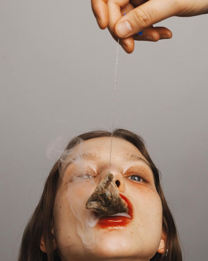 Conceptual beauty portrait of a woman with a teabag on her nose, red lipstick, and an outstretched hand holding a string.