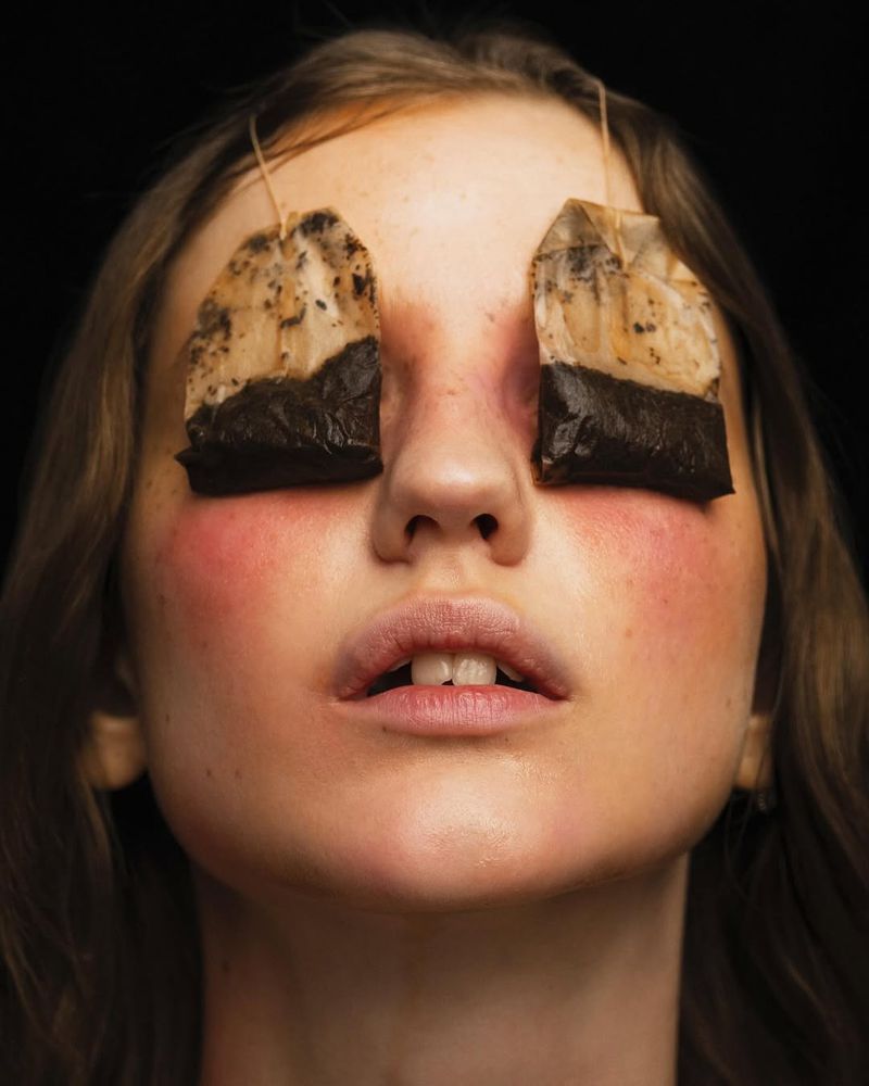 Close-up portrait of a young woman with tea bags covering her eyes, set against a dark backdrop. The image emphasizes skincare or beauty.