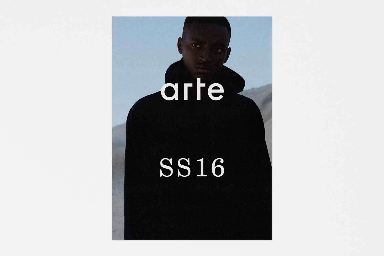 Portrait of a Black man wearing a black hoodie with overlaid white text reading arte SS16 against a pale sky background