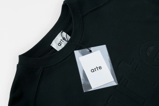 Close-up of a black Arte sweatshirt with a hanging paper brand tag and collar label against a white seamless background