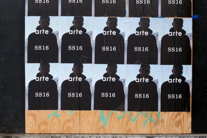 Grid of repeated Arte SS16 posters wheat-pasted on a wooden construction wall with white typography.