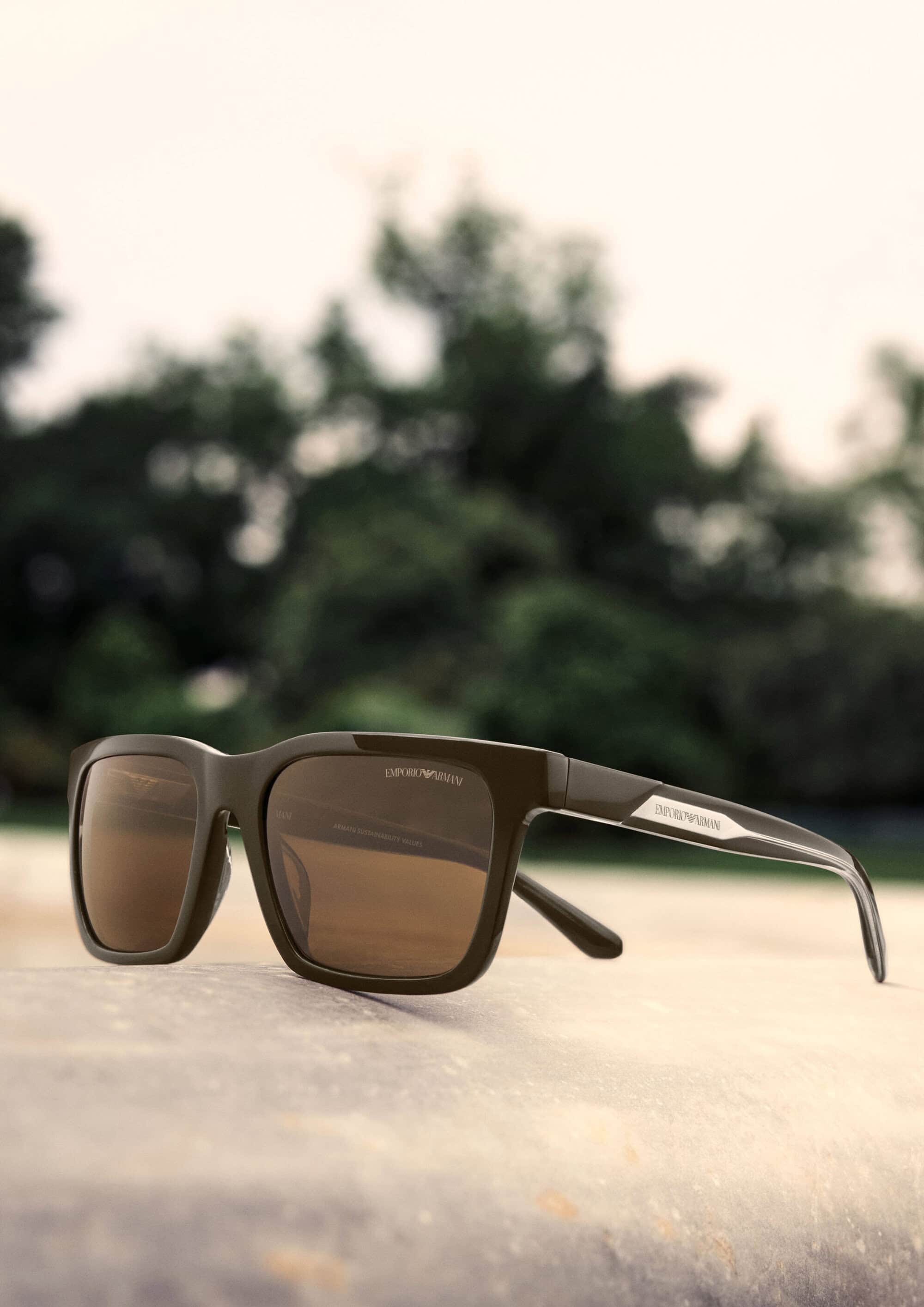 Emporio Armani dark brown sunglasses resting on a light surface outdoors with a blurred green park background.