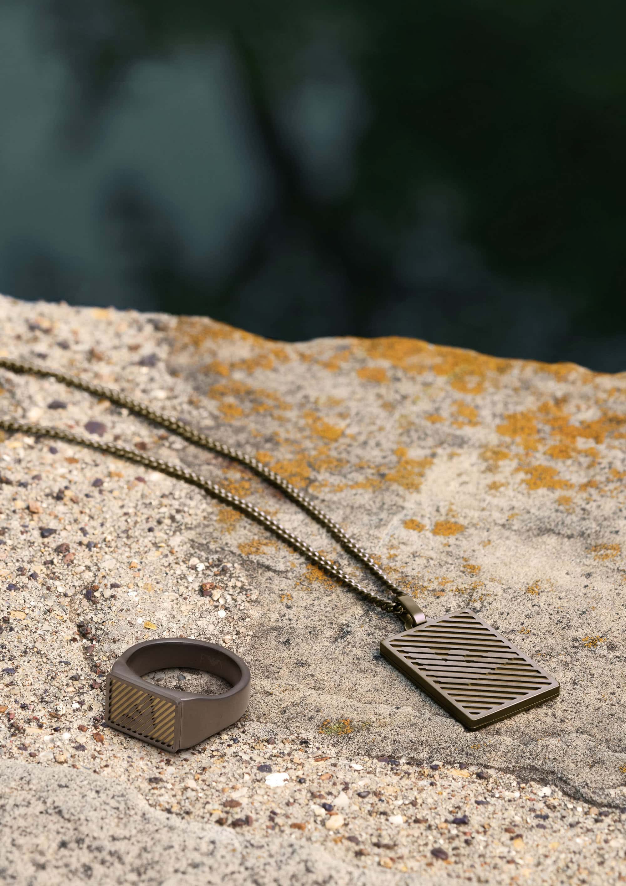 Close-up of a bronze metal ring and matching rectangular pendant necklace resting on a weathered stone surface outdoors.