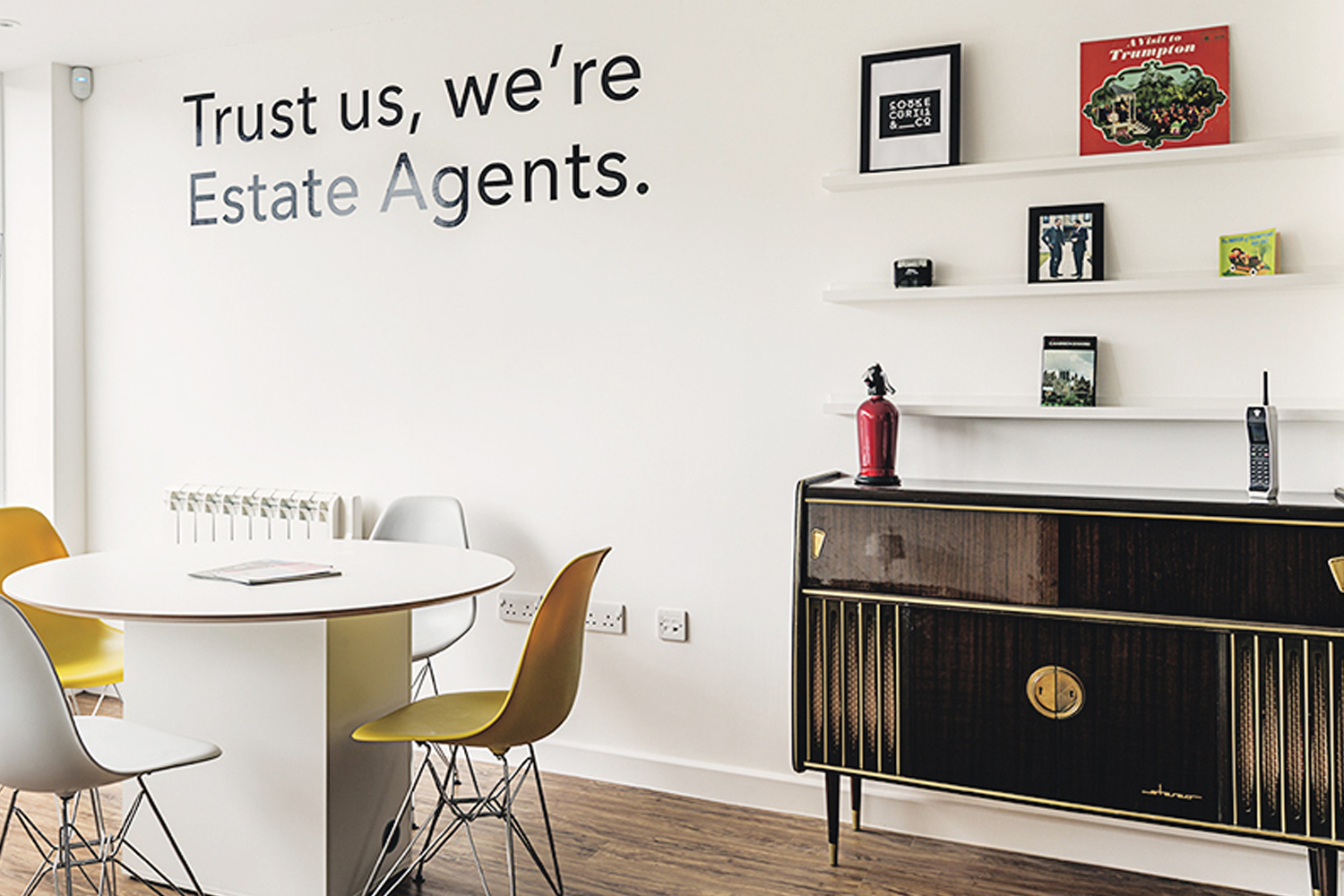 Interior of an office featuring a white round table, yellow and white Eames-style chairs, and wall text reading Trust us we are Estate Agents.