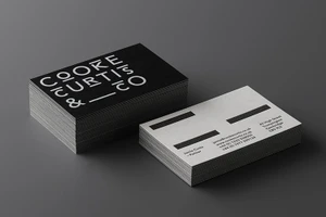 Two stacks of business cards for Cooke Curtis and Co on a dark grey surface, showing black and white minimalist typography.