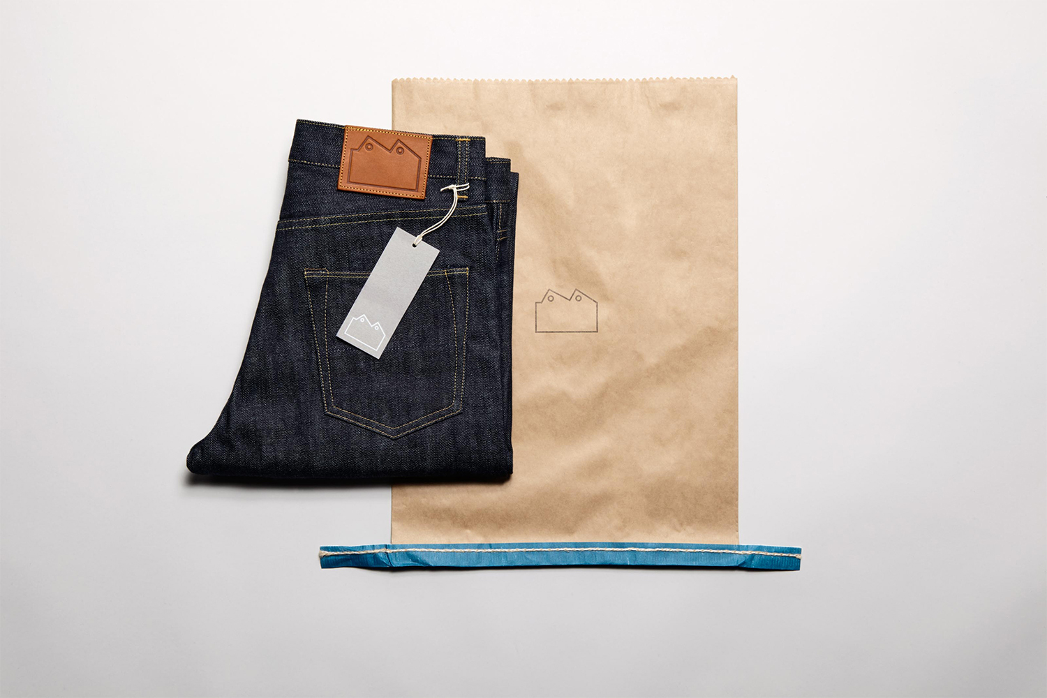 Folded raw denim jeans with a brown leather patch and hang tag laid next to a brown paper bag on white background
