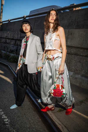 Two models standing near a stone wall wearing Kenzo designer fashion clothing with floral patterns and holding a patterned handbag.
