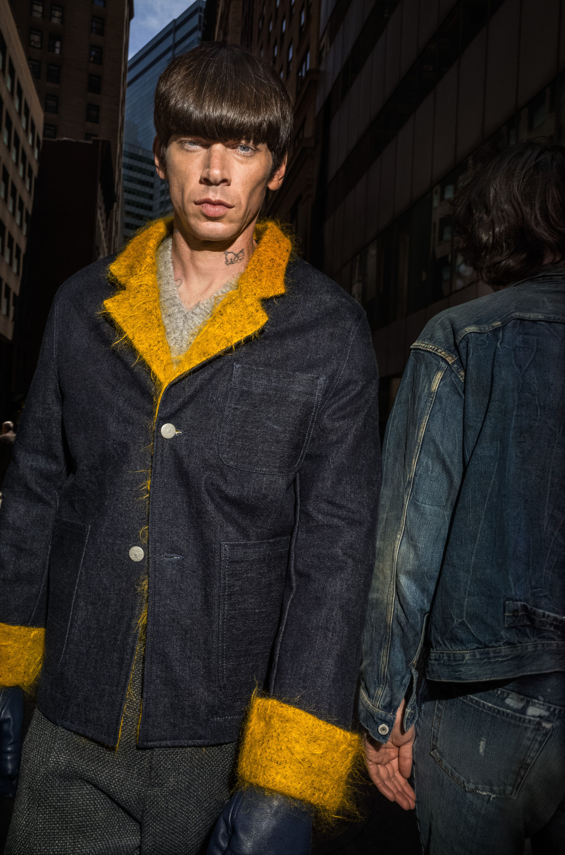 Model with bowl haircut wearing a dark denim jacket with bright yellow fluffy trim on an urban street.
