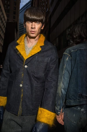 Model with bowl haircut wearing a dark denim jacket with bright yellow fluffy trim on an urban street.