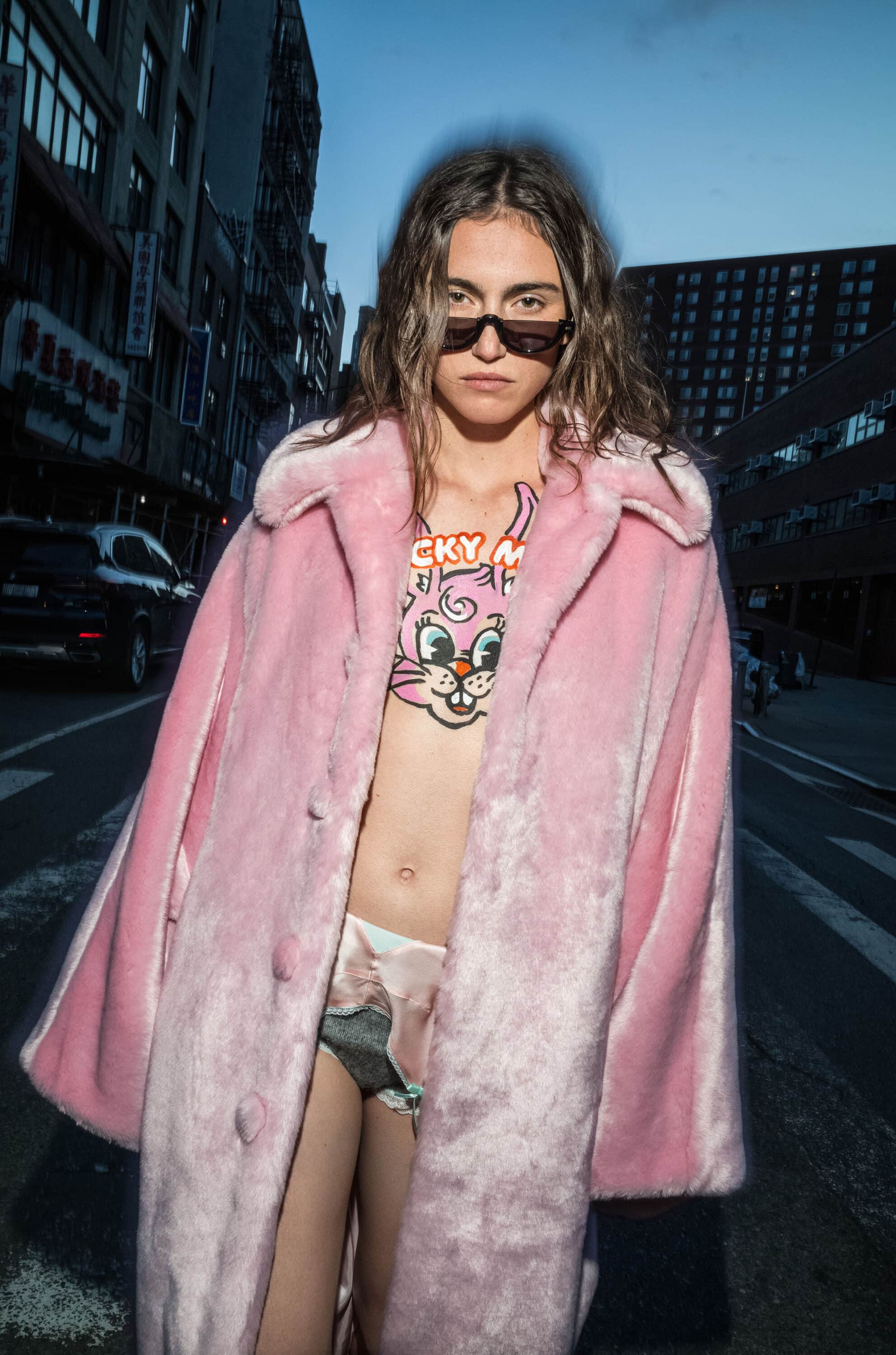 Model wearing a pink fur coat and dark sunglasses standing on a city street at dusk for Kenzo fall 2025 ad campaign.
