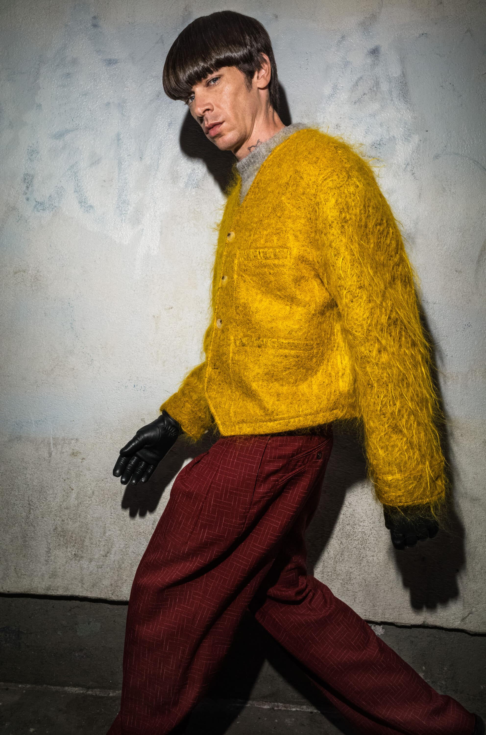 Model in a fuzzy yellow cardigan and red trousers wearing black gloves against a textured concrete wall.