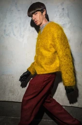 Model in a fuzzy yellow cardigan and red trousers wearing black gloves against a textured concrete wall.