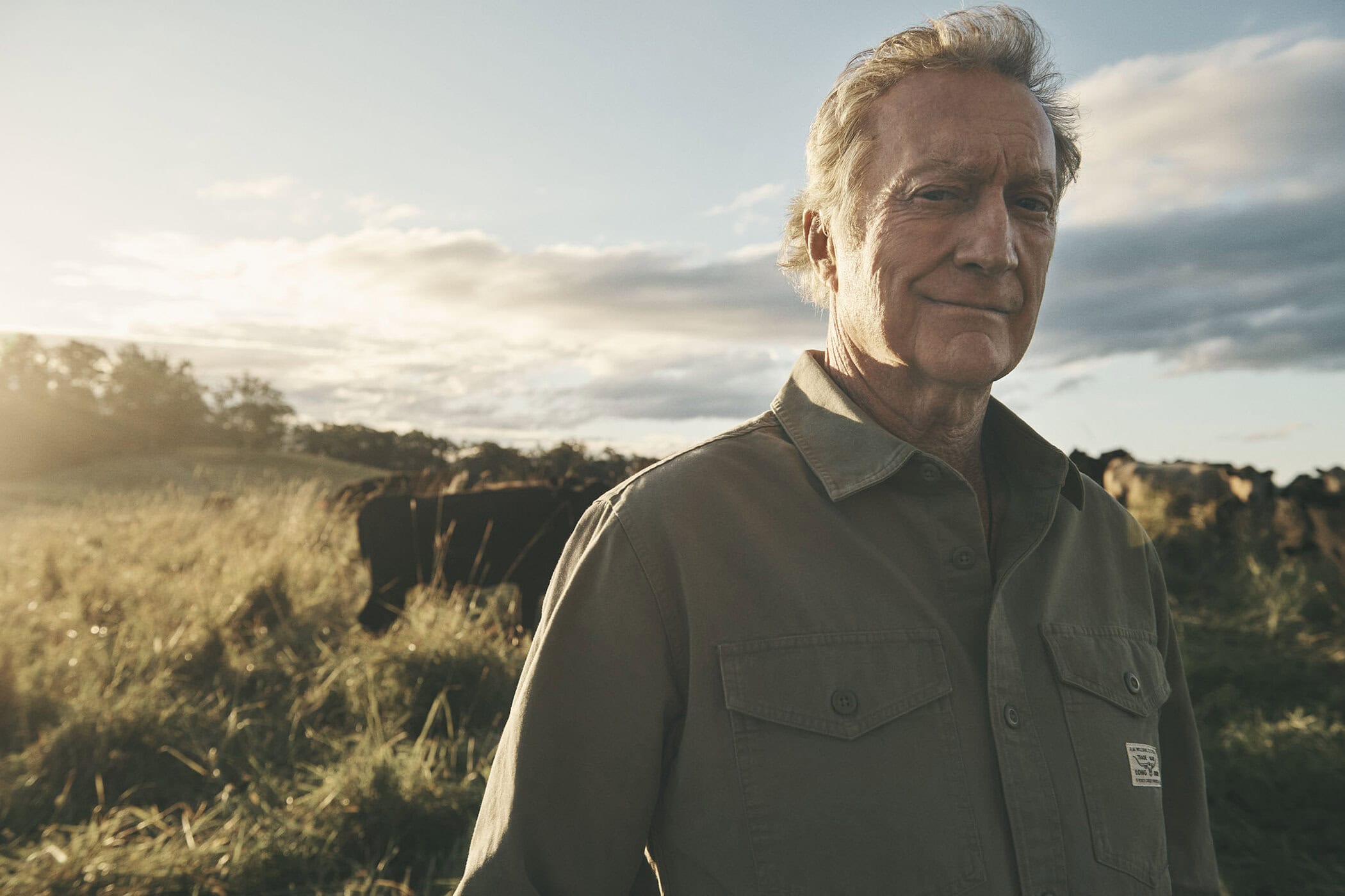 An older man wearing an olive green button-up shirt standing in an open grassy field under a cloudy sunset sky.