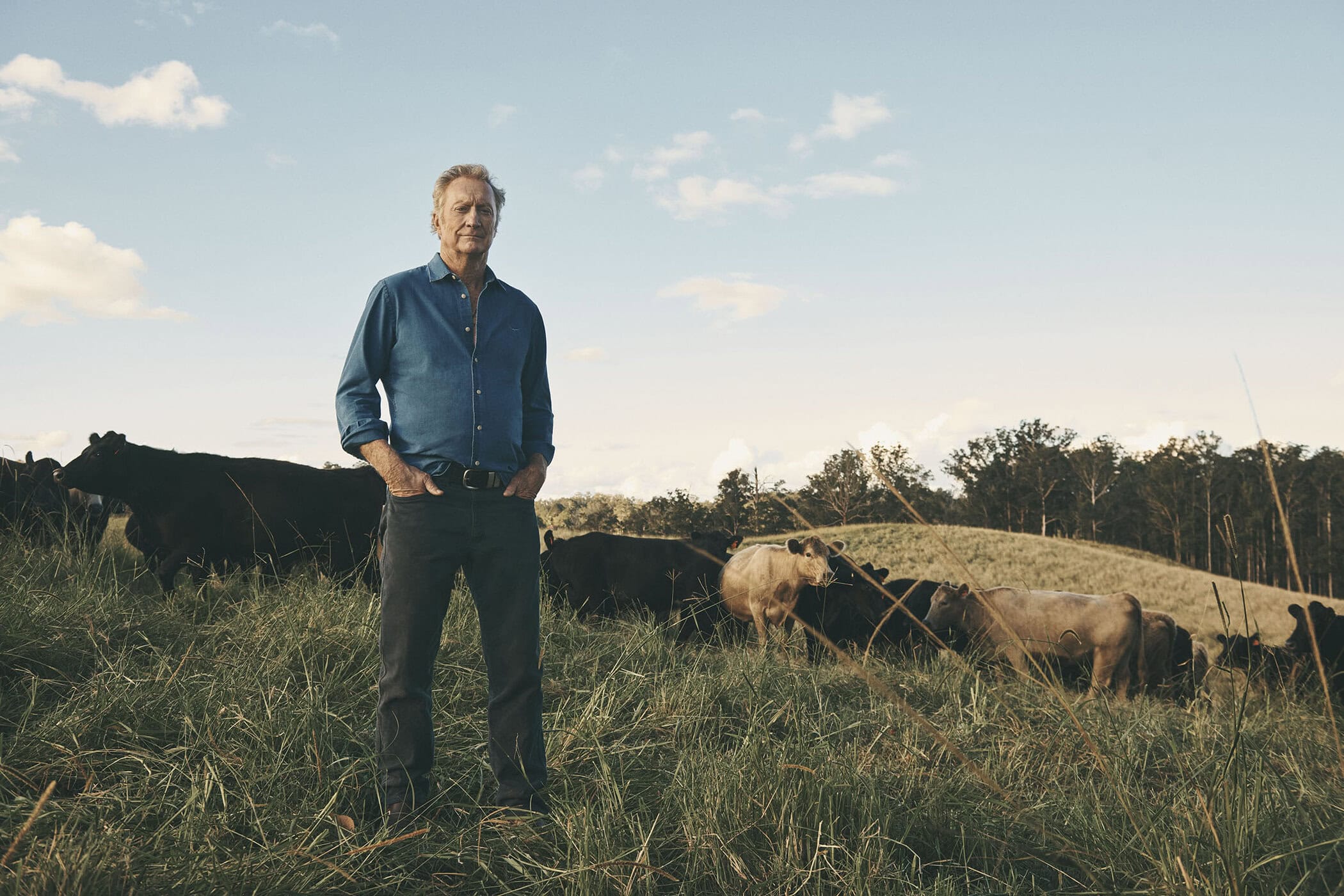 An older man standing in a grassy field with cattle under an open sky for R.M. Williams