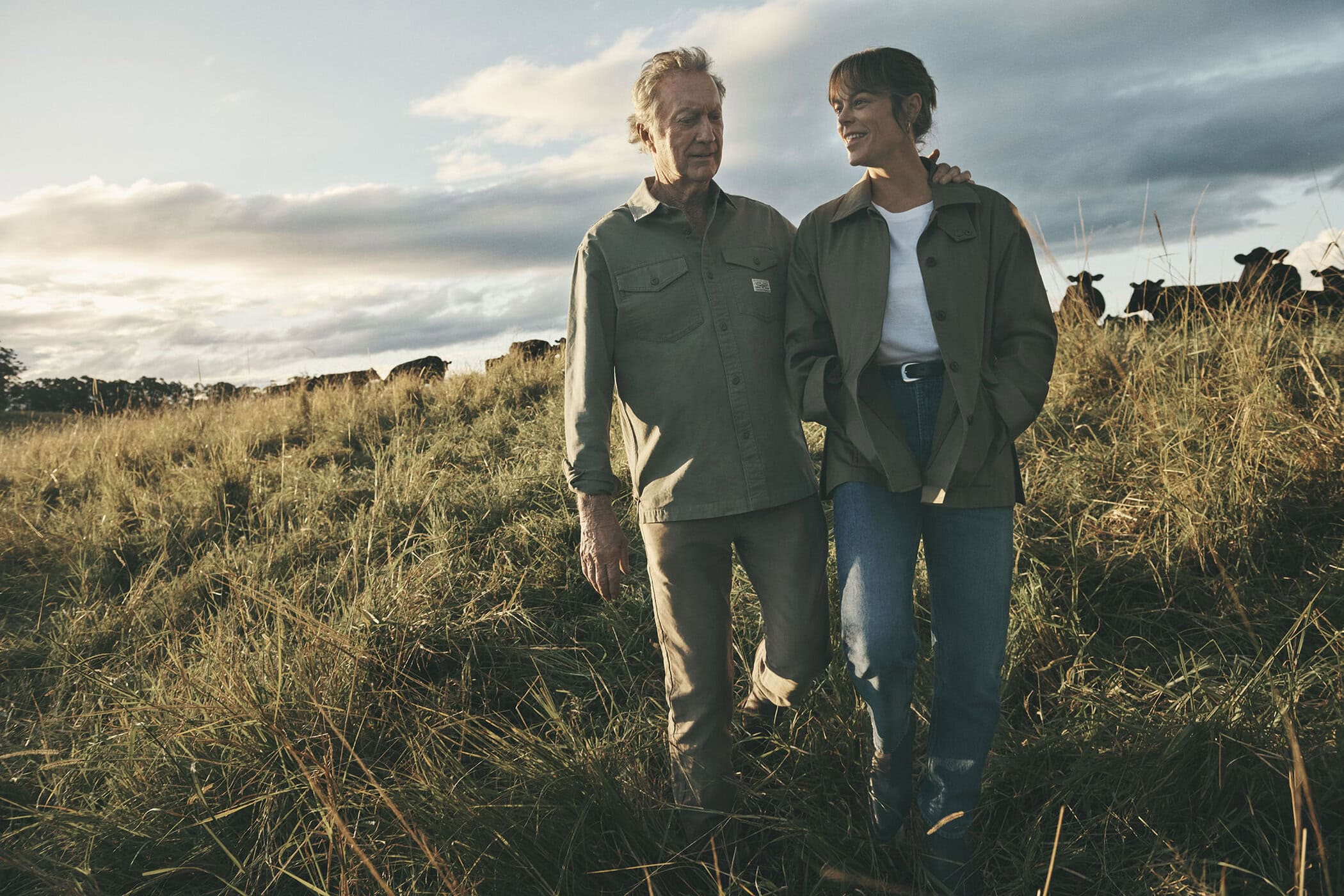 An older man and a younger woman walking together through a tall grass meadow under a cloudy sky for R.M. Williams
