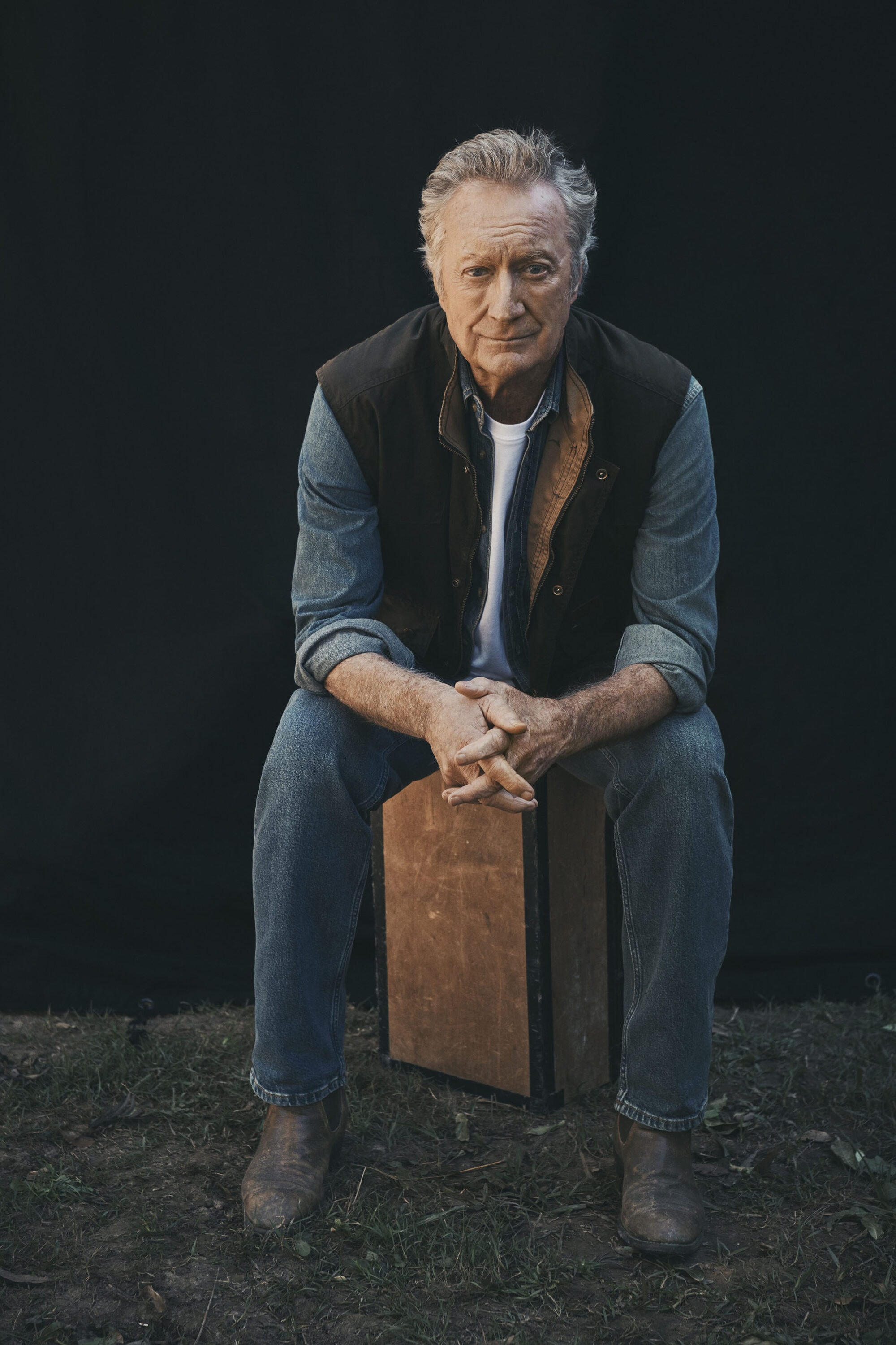 Bryan Brown seated on a wooden box wearing denim jeans, a white t-shirt, denim shirt, and dark vest against a dark backdrop