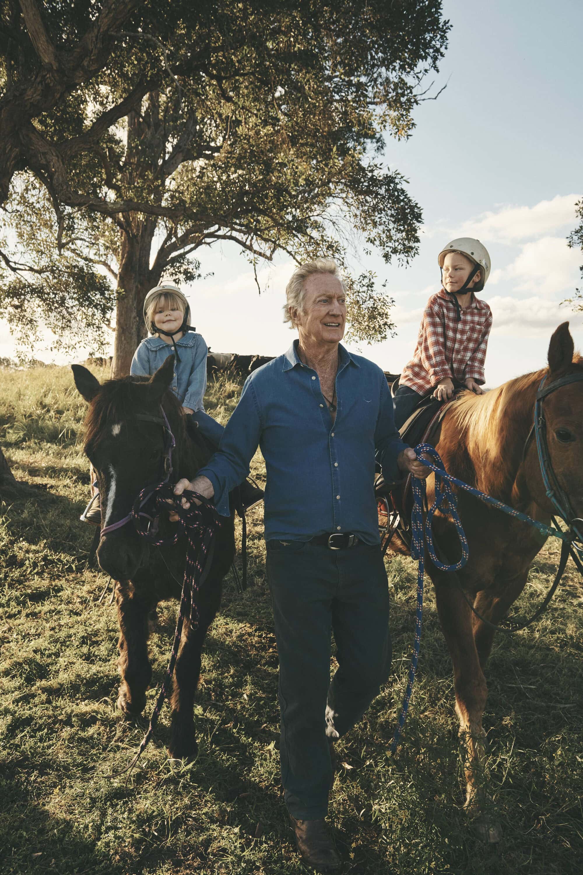 Bryan Brown walking between two horses with children mounted, outdoors under trees in a grassy field