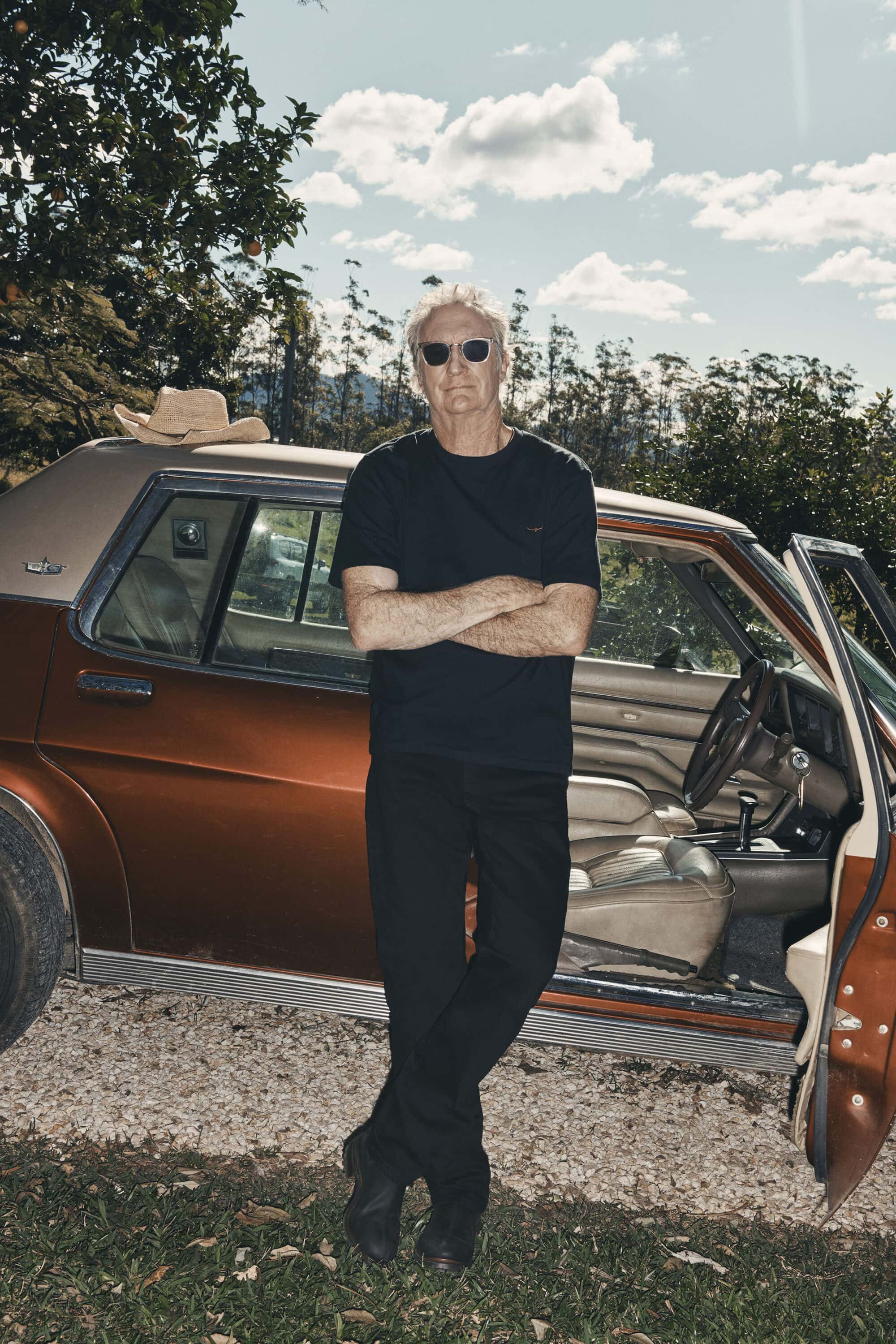 Man wearing sunglasses and black t-shirt standing with arms crossed beside a brown car outdoors in Australia.