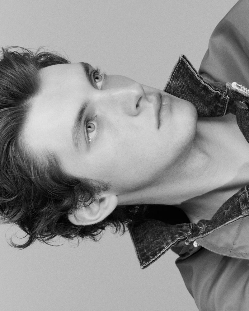 Portrait of a young fair-skinned adult male with dark hair, gazing upward in a close-up shot wearing a denim collar jacket, black and white.