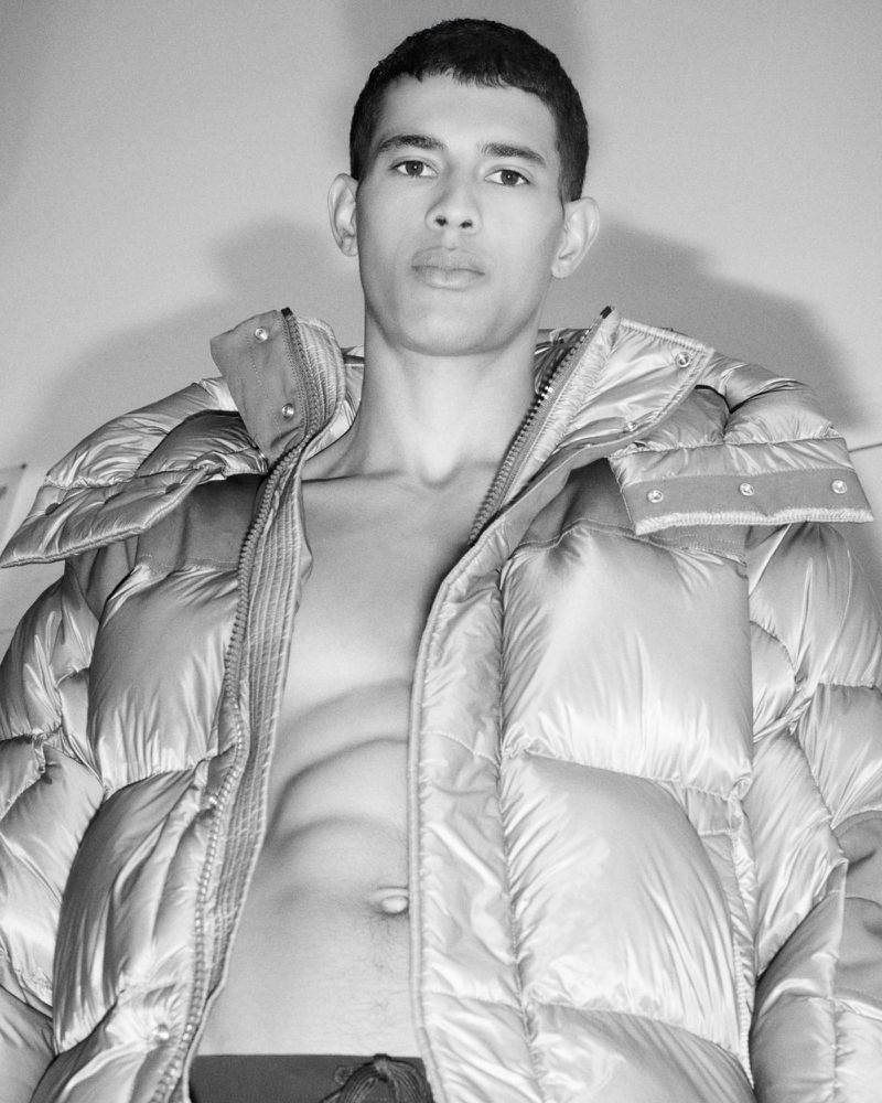 Monochrome portrait of adult male wearing puffy jacket open, fair skin, looking at camera. The background is soft neutral tone.
