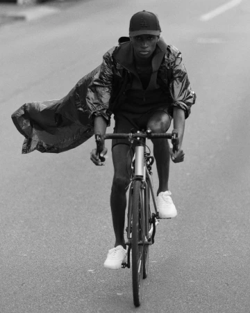 A black and white shot captures a person riding a bicycle, wearing athletic wear, a cap, and a jacket that blows in the wind.