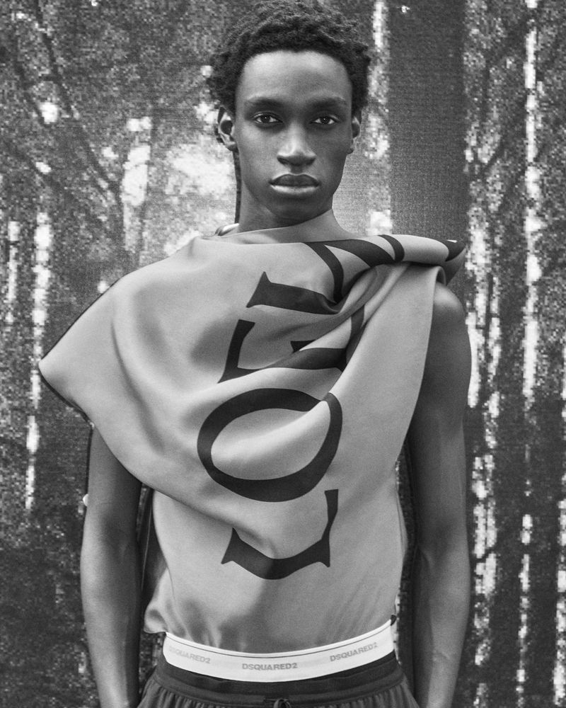 Portrait of a slender Black male model wearing a draped printed top, posing against a textured monochrome forest background in a studio.