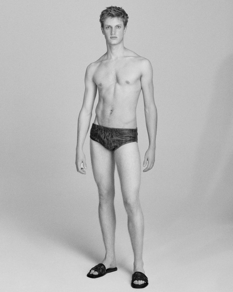 Full-length black and white studio shot of a shirtless adult male in swim trunks and sandals, standing in a neutral, gray background.