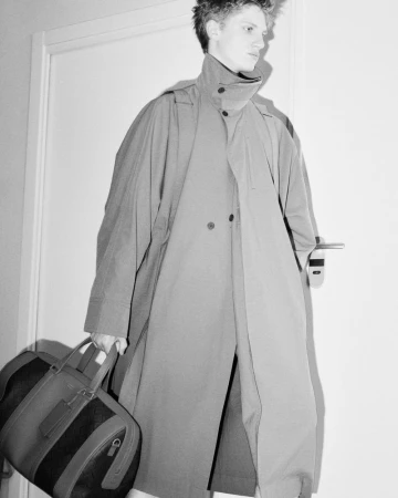 Monochrome medium shot of a fair-skinned young adult in a coat carrying a duffel bag in an indoor setting with a neutral wall.